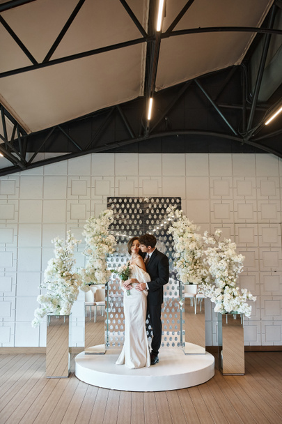 повна довжина нареченого, що обіймає чарівну жінку з весільним букетом біля квіткового декору в банкетному залі - Фото, зображення