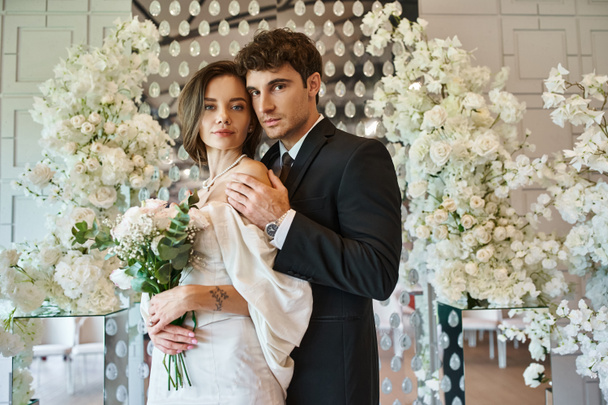 giovani ed eleganti sposi guardando la fotocamera vicino composizione floreale bianca in luogo di nozze - Foto, immagini