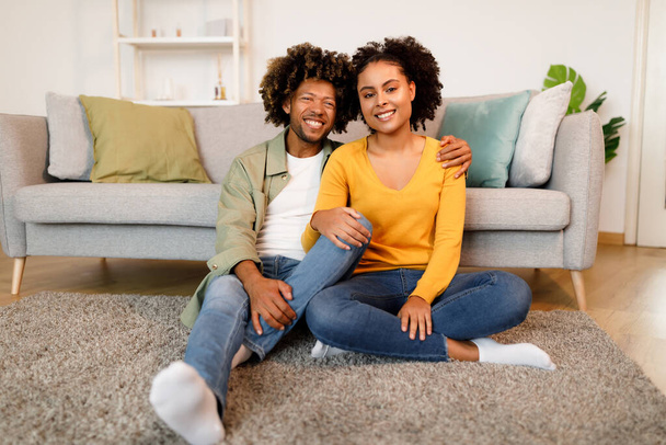 Ρομαντικές αφρικανικές αμερικανικές συζύγους αγκαλιάζει κάθεται στο πάτωμα κοντά στον καναπέ στο σπίτι, εκφράζοντας θετικά συναισθήματα χαμογελώντας στην κάμερα στο σύγχρονο εσωτερικό του σαλονιού. Ο σύζυγος αγκαλιάζει τη νεαρή σύζυγό του - Φωτογραφία, εικόνα