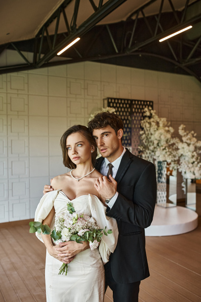 elegantní ženich objímající půvabnou nevěstu se svatební kyticí a při pohledu na kameru v sále - Fotografie, Obrázek