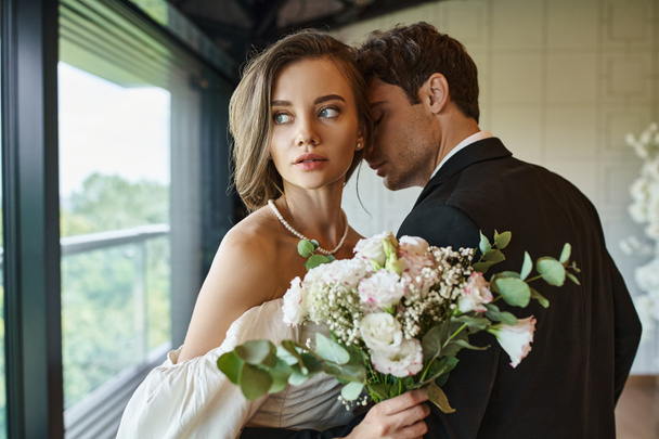 okouzlující nevěsta se svatební kyticí dívá pryč v blízkosti ženicha v černém obleku v hodovní síni - Fotografie, Obrázek
