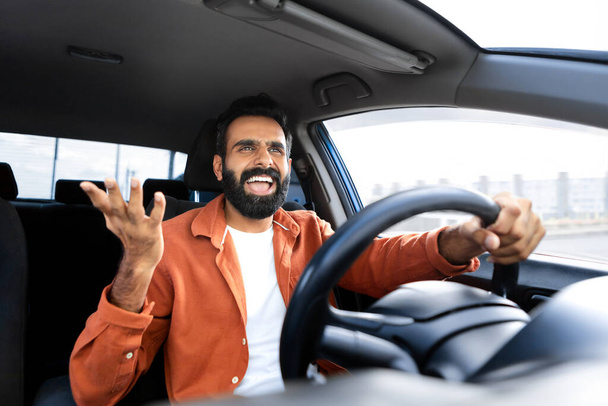 Безумный агрессивный индиец, кричащий, сидя в кресле водителя и держа руль внутри современного авто, парень, управляющий опасным выражением недовольства. Проблема пробок, риск ДТП - Фото, изображение