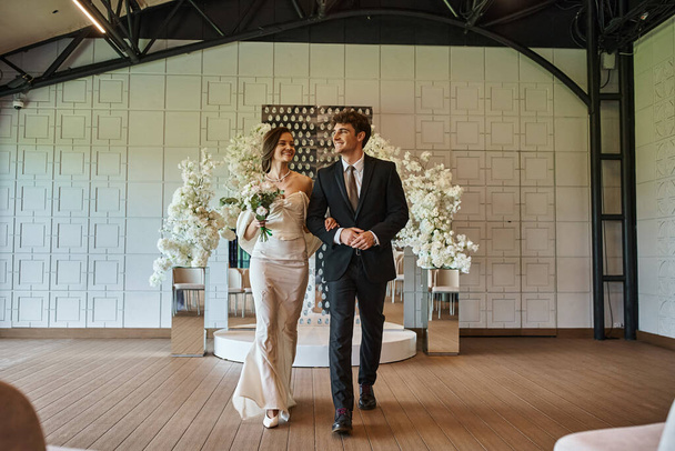 volledige lengte van elegante vrolijke paar in bruiloft kleding wandelen in evenementenhal met bloemen decor - Foto, afbeelding