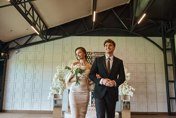 alegre pareja elegante en traje de novia caminando en la sala de eventos moderna con decoración floral blanca - Foto, Imagen