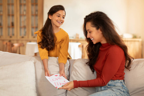 Mothers Day Surprise. Cheerful Arabic Preteen Kid Daughter Congratulating Mom On Holiday Or Birthday, Giving Handmade Greeting Card Painting To Her, Celebrating In Modern Living Room At Home - Photo, Image