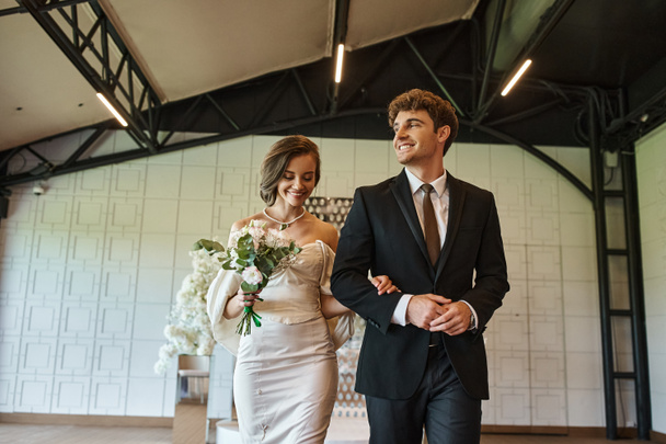 happy bride in white dress and groom in black suit smiling in modern decorated wedding venue - Photo, Image