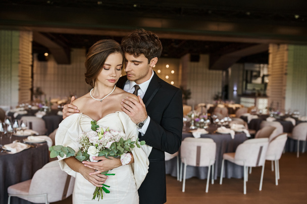elegante novio con los ojos cerrados abrazando encantadora novia con ramo de boda en la sala de eventos moderna - Foto, Imagen