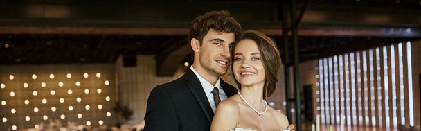 giovane e gioiosa coppia di sposi nel sorridere alla macchina fotografica in sala celebrazione decorata, striscione - Foto, immagini