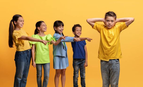 Angry cruel multiracial kids teenagers laughing and pointing at their classmate boy covering his ears with hands. School aged children bullying child. Social inequality problem. Schoolchildren baiting - Photo, Image