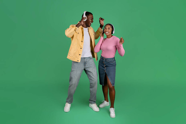 Cheerful african american millennial man and woman using wireless headphones isolated on green background. Happy young black couple listening to music and dancing together, copy space, full length - Photo, Image
