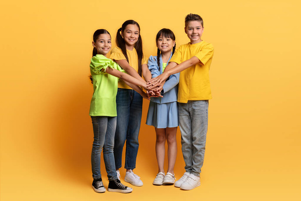 Happy multiethnic teenagers holding hands on top of each other and smiling at camera. Cheerful kids boys and girls have fun together isolated on yellow background, playing game. Happy childhood - Photo, Image