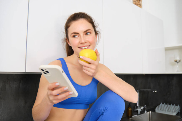 Portrait of sportswoman, girl eating and apple and looking at her social media, smartphone screen, having a snack in kitchen, wearing fitness activewear. - Photo, Image