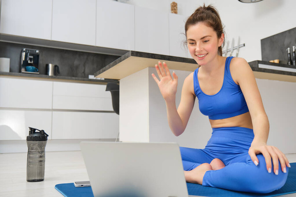 Портрет інструктора фітнесу, жінка підключається до онлайн-тренувальної сесії, хвилі рука в ноутбуці, навчає тренування йоги з дому, сидить на гумовому килимку. - Фото, зображення