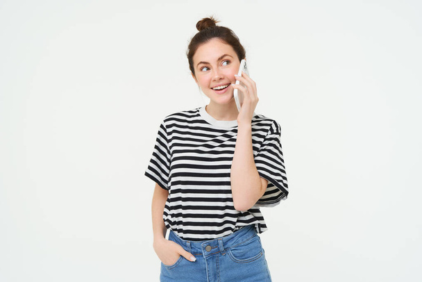 Fröhliche junge Frau telefoniert mit dem Handy, chattet am Telefon, nutzt das Smartphone, steht vor weißem Hintergrund. Kopierraum - Foto, Bild