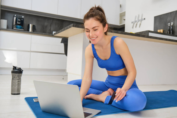 Portret młodej kobiety po lekcji fitness online w domu, siedzącej na macie do jogi i korzystającej z laptopa podczas treningu aerobiku. - Zdjęcie, obraz