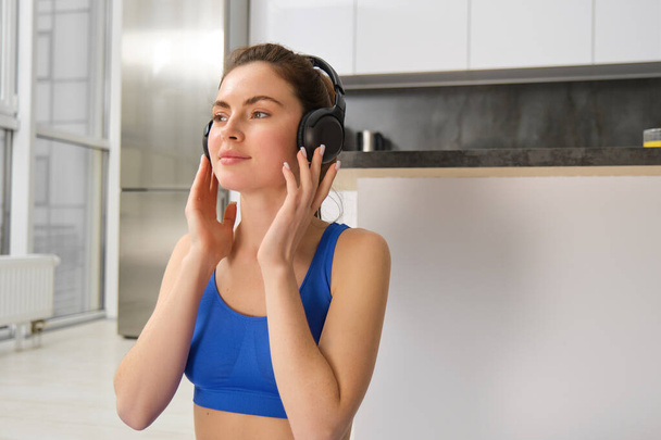 Jonge vrouw zet een draadloze hoofdtelefoon op om muziek te luisteren tijdens de training thuis, draagt een sportbeha, glimlacht met een blij gezicht. - Foto, afbeelding