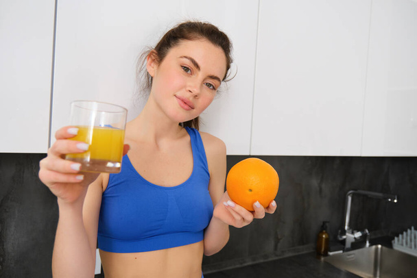 Close up portrait of sportswoman, fitness girl holding glass of fresh juice and an orange in hands, smiling at camera, standing in kitchen. Workout and healthy lifestyle concept - Photo, Image