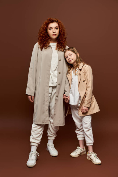 two generations, curly mother holding hands with daughter and standing in coats on brown backdrop - Foto, Bild