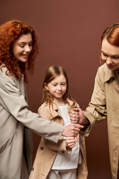 generaciones, familia feliz con el pelo rojo que apilan las manos juntas en el telón de fondo marrón, mujeres y niñas - Foto, imagen