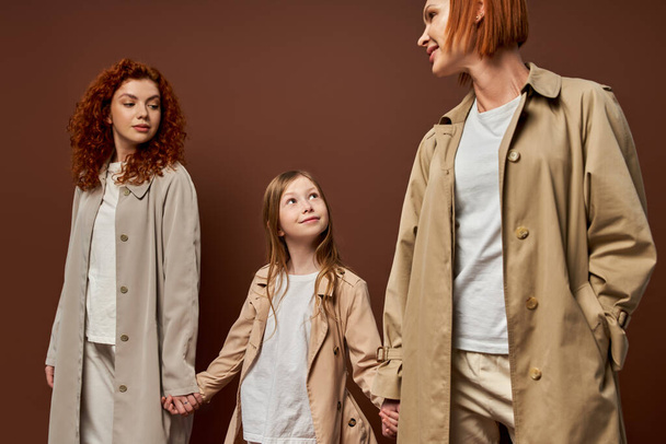 Üç nesildir, paltolu kadınlar kahverengi arka planda mutlu bir kızla el ele tutuşuyorlar, kızıl saçlı bir aile. - Fotoğraf, Görsel