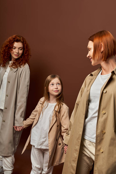 három generáció, boldog vörös hajú család kabátban kéz a kézben barna háttér, nők és lány - Fotó, kép