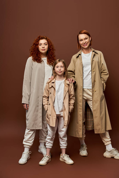 female generation, redhead family standing together in outerwear on brown backdrop, women and girl - Fotó, kép