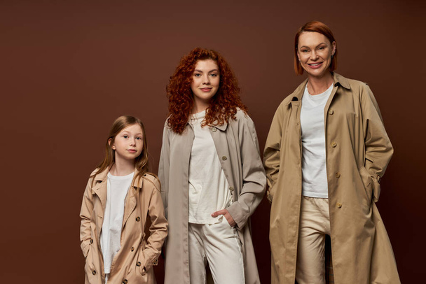 generación femenina, familia con el pelo rojo posando con las manos en bolsillos en ropa de abrigo sobre fondo marrón - Foto, Imagen