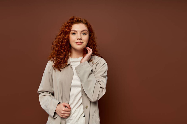 portrait of pleased woman with red curly hair looking at camera on brown background, autumn colors - Photo, Image