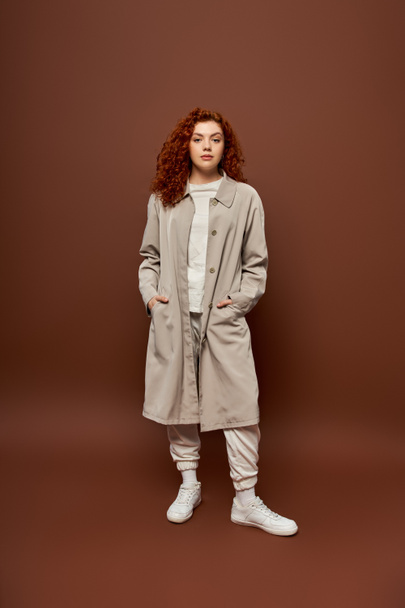 curly woman with red hair posing in autumn coat and joggers on brown background, hands in pockets - Photo, Image