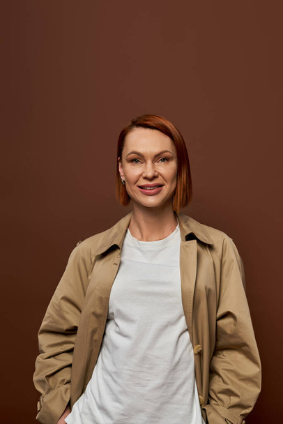 joyful redhead woman in beige trench coat looking at camera on brown backdrop, autumn fashion - Photo, Image