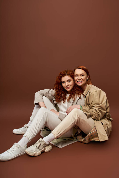 два покоління, позитивні жінки з рудим волоссям позують в осінніх траншеях пальто на коричневому фоні - Фото, зображення
