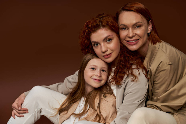 three generations of women, happy redhead family looking at camera and smiling on brown background - Photo, Image