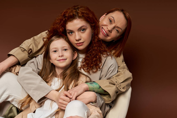 three generations, positive redhead family looking at camera on brown background, modern parenting - Photo, Image
