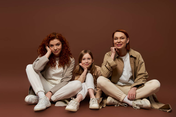 happy redhead family of three female generations sitting on brown background, autumn coats - Foto, imagen