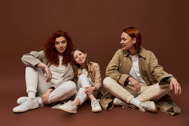 cheerful redhead family of three female generations sitting on brown background, autumn coats - Foto, Imagem