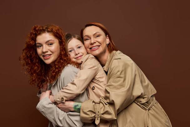 happy redhead family of three female generations hugging on brown background, autumn fashion - Фото, изображение