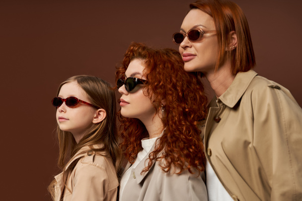three female generations with red hair posing in sunglasses and coats on brown backdrop, family bond - Φωτογραφία, εικόνα
