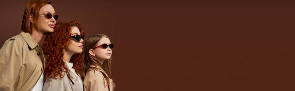three female generations with red hair posing in sunglasses and coats on brown backdrop, banner - Fotografie, Obrázek