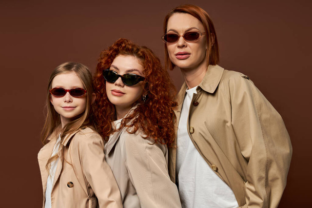 three female generations with red hair posing in sunglasses on brown backdrop, hands in pockets - Foto, Bild