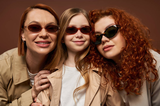 conceito de mudança de geração familiar, mulheres ruivas felizes e menina em óculos de sol no fundo marrom - Foto, Imagem