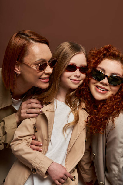 family generation change concept, cheerful redhead women and girl in sunglasses on brown backdrop - Foto, Bild