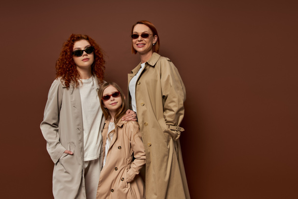 family portrait of female generations in sunglasses and coats on brown background, hands in pockets - Foto, imagen