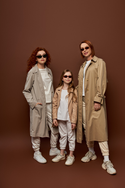 family photo of female generations in sunglasses and coats posing on brown background, full length - Fotó, kép
