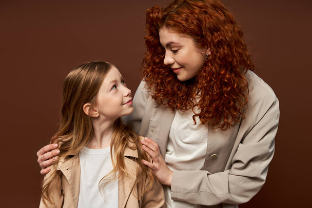 два покоління, молода кучерява мати з рудим волоссям дивиться на дочку на коричневому фоні, сім'я - Фото, зображення