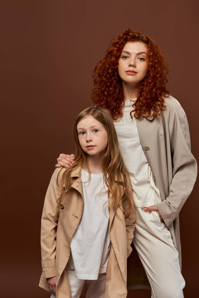 two generations, young curly mother looking at camera with redhead daughter on brown backdrop - Photo, Image