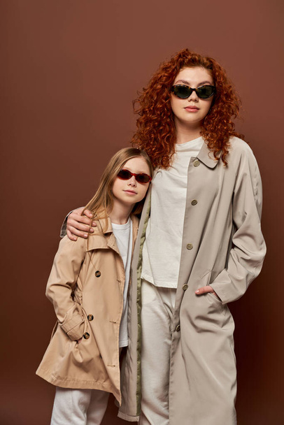 autumn style, redhead mother and kid posing in sunglasses and beige trench coats, brown backdrop - Photo, Image