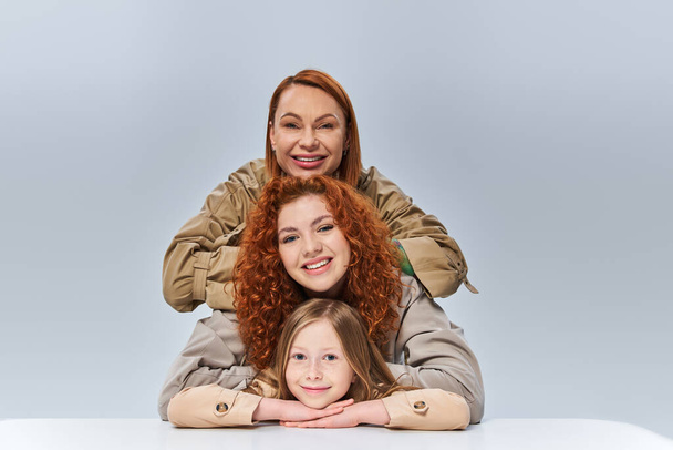 three generations, redhead mother and child in beige trench coats posing on grey background - Fotografie, Obrázek