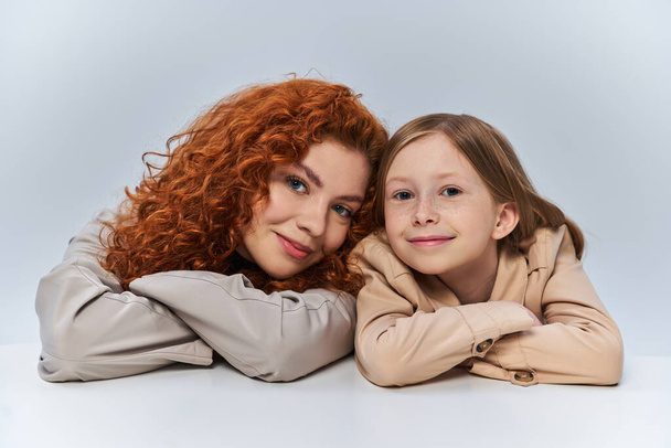 boldog két generáció, vörös hajú anya és gyermek bézs ballonkabátban mosolygós szürke háttér - Fotó, kép