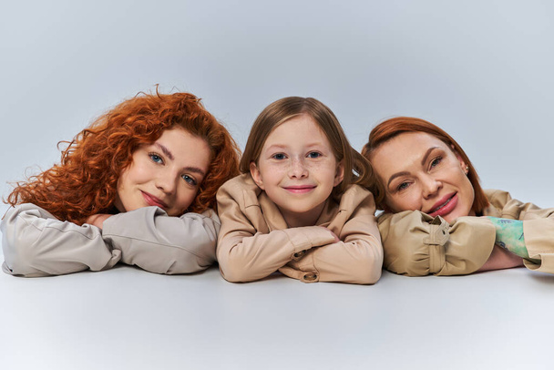 gelukkig vrouwelijke generaties, roodharige vrouwen en kind in beige jassen glimlachen op grijze achtergrond, familie - Foto, afbeelding