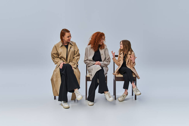 three generations, redhead girl talking to her relatives and sitting on chairs on grey backdrop - Photo, Image